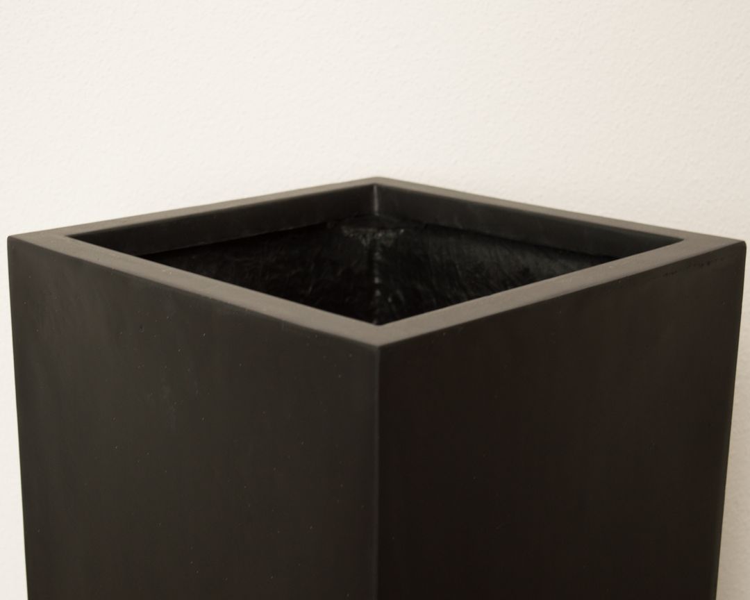 Blick von schräg oben auf die quadratische Öffnung eines schwarz-matten Säulen-Pflanzkübels aus Fiberglas vor heller Wand