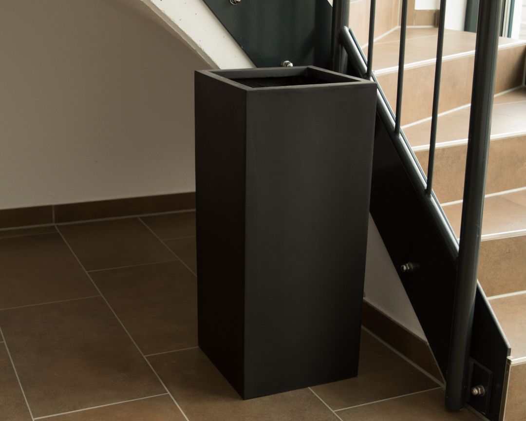 Schwarz-matter Pflanzkübel aus Fiberglas in Säulenform ohne Bepflanzung neben einer Treppe auf Fliesenboden