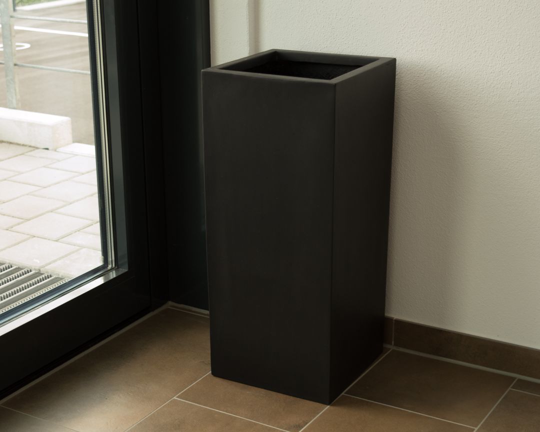Schwarz-matter quadratischer Pflanzkübel aus Fiberglas in Säulenform, leer neben einer Glastür auf braunem Fliesenboden
