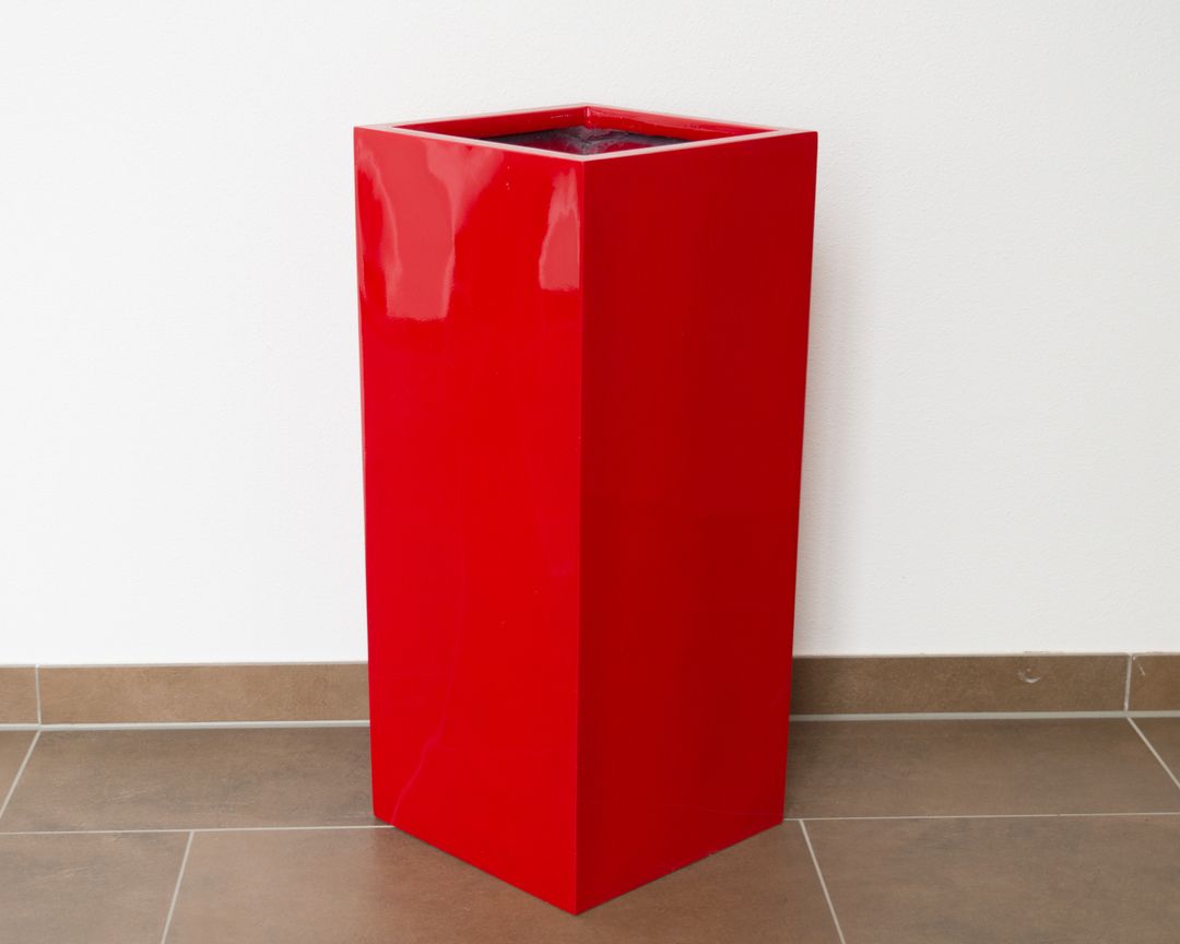 Rote quadratische Pflanzkübel-Säule aus Fiberglas in Hochglanz, leer vor weißer Wand auf braunem Fliesenboden