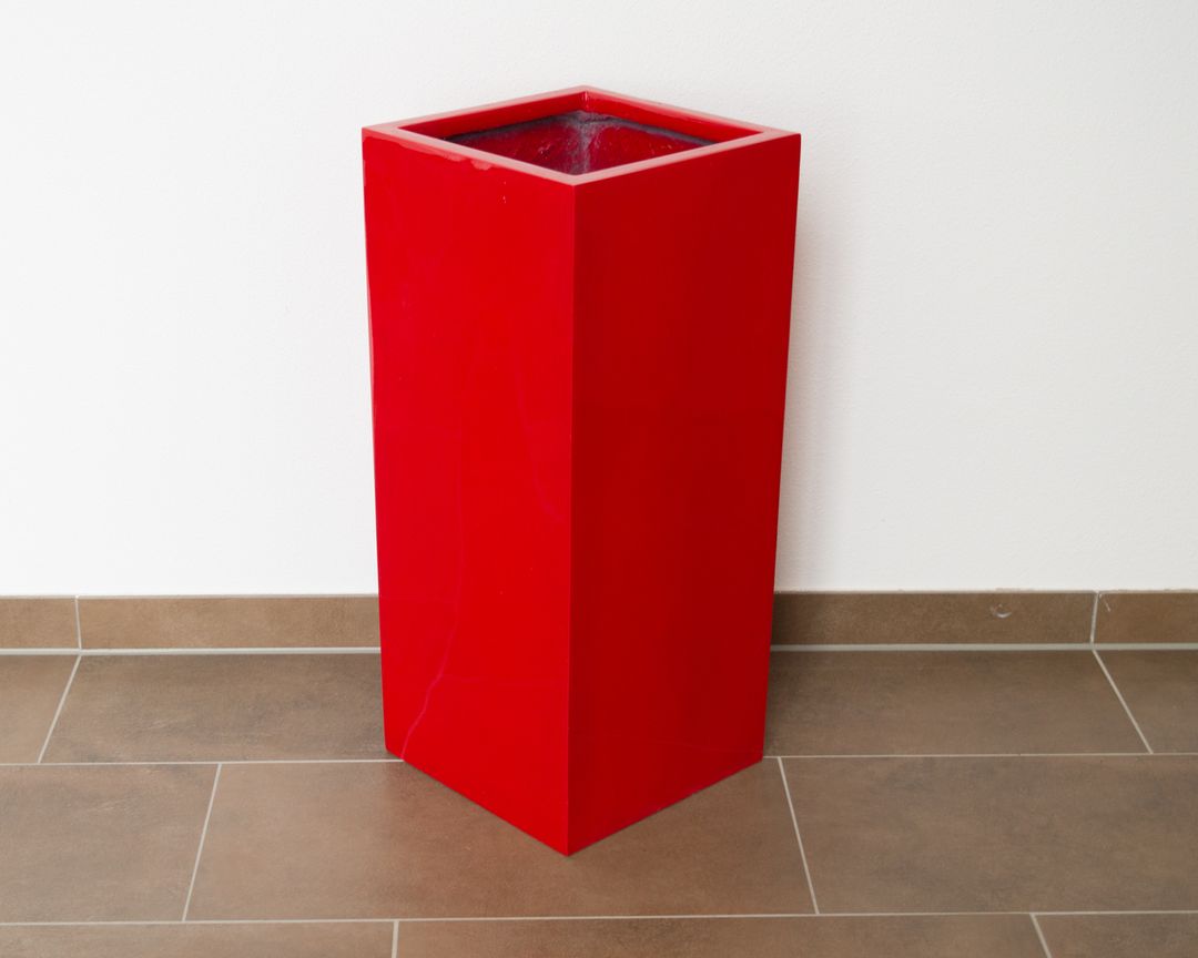Hochglanz roter Pflanzkübel aus Fiberglas in Säulenform, leer auf braunem Fliesenboden vor weißer Wand