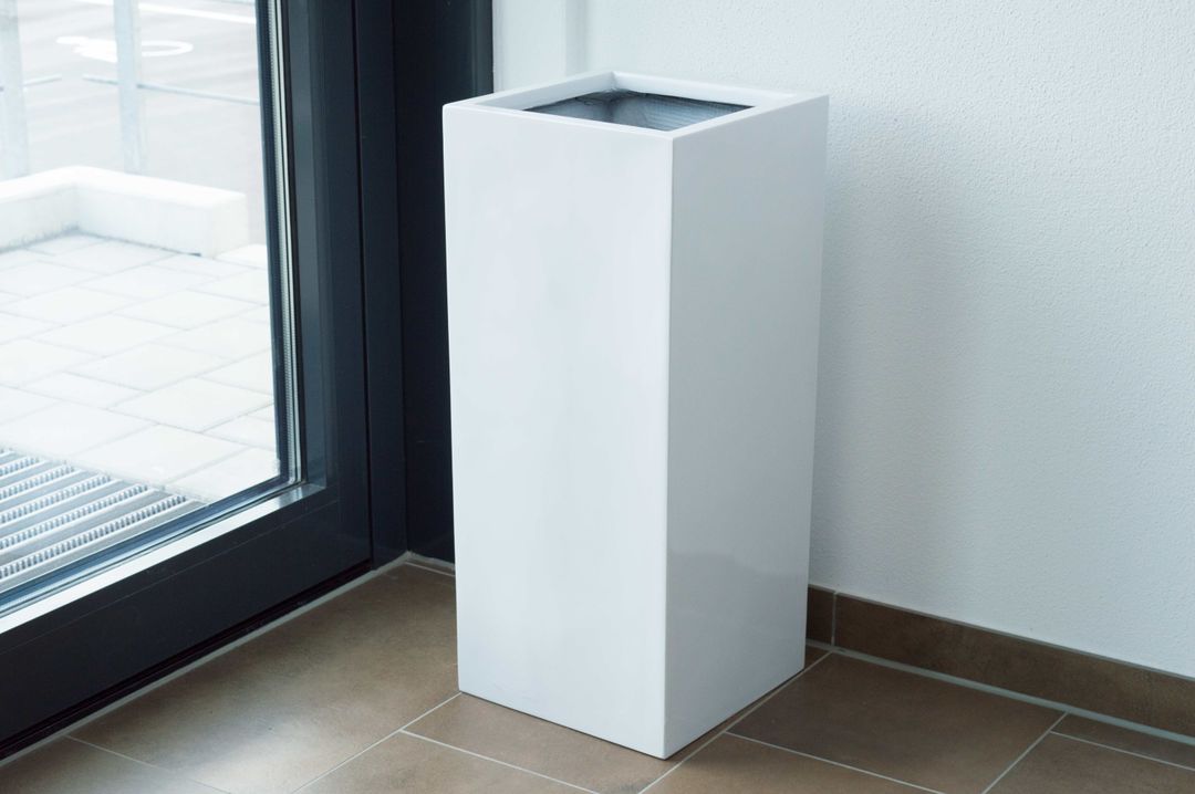 Weißer Hochglanz-Pflanzkübel in Säulenform ohne Bepflanzung neben einer Glastür auf braunem Fliesenboden