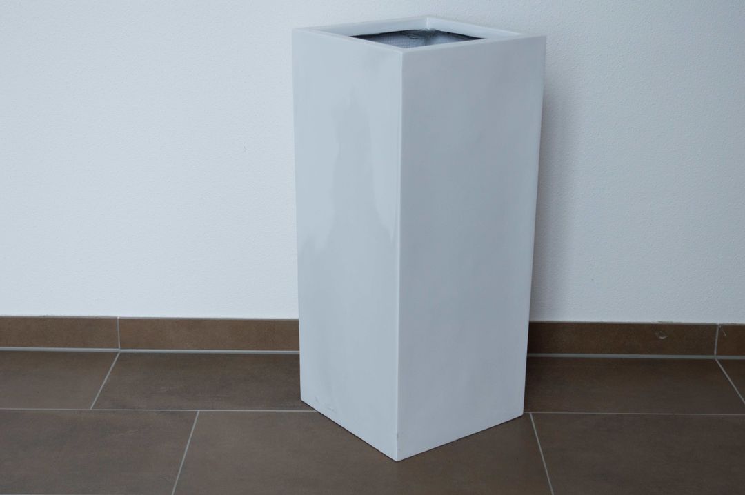 Hochglanz weißer Pflanzkübel aus Fiberglas in Säulenform, leer vor weißer Wand auf braunem Fliesenboden