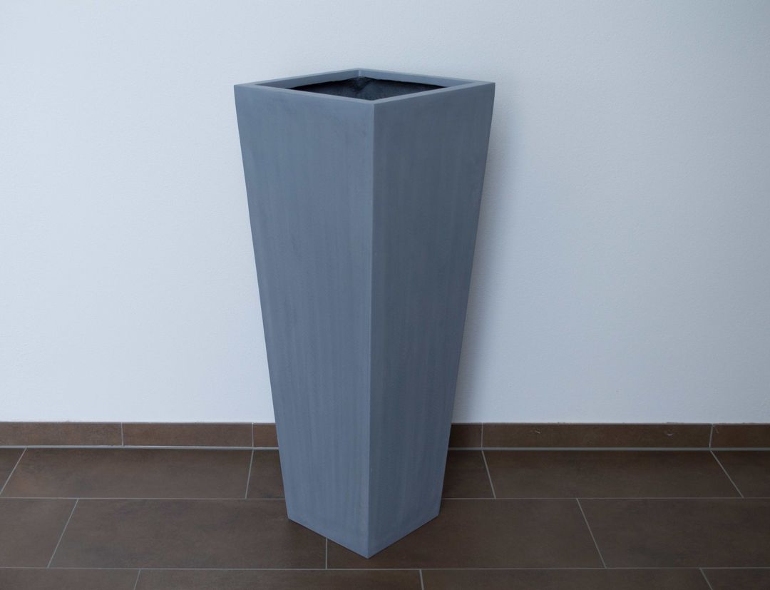 Grauer konischer Pflanzkübel aus Fiberglas in Säulenform ohne Bepflanzung vor weißer Wand auf braunem Fliesenboden