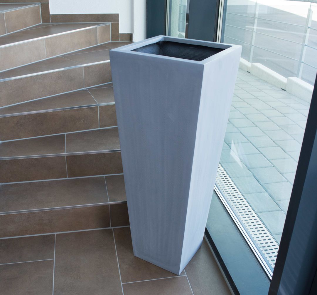 Grauer konischer Pflanzkübel aus Fiberglas in Säulenform, leer, neben einer gefliesten Treppe vor einer Glastür