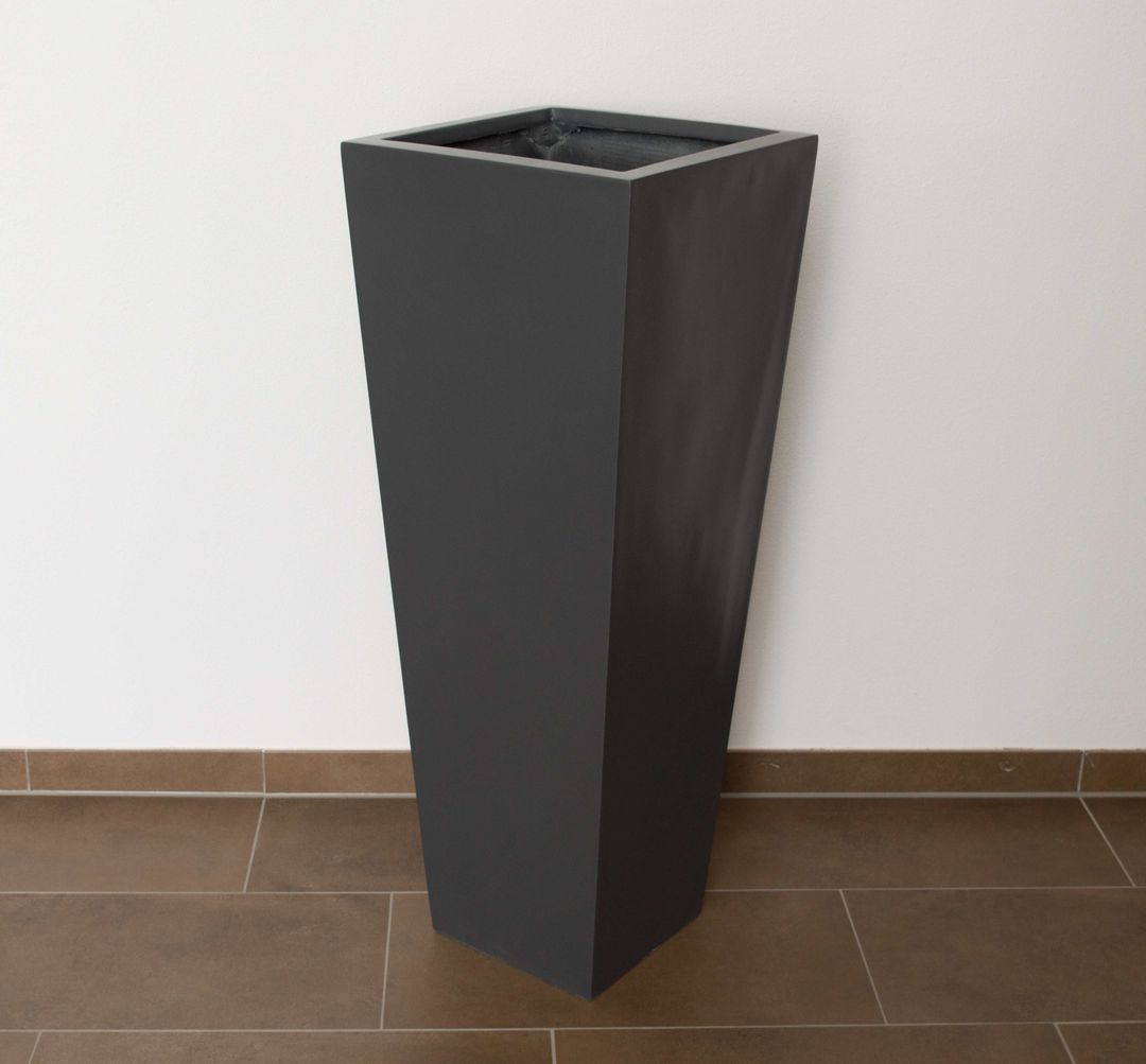 Konischer Pflanzkübel aus Fiberglas in Anthrazit metallic als hohe Säule vor weißer Wand auf braunem Fliesenboden