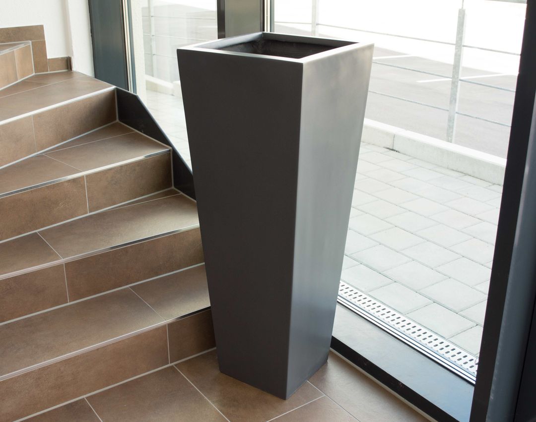 Hohe, konische Pflanzkübel-Säule aus Fiberglas in Anthrazit metallic, leer im Eingangsbereich neben Treppe und Glasfront