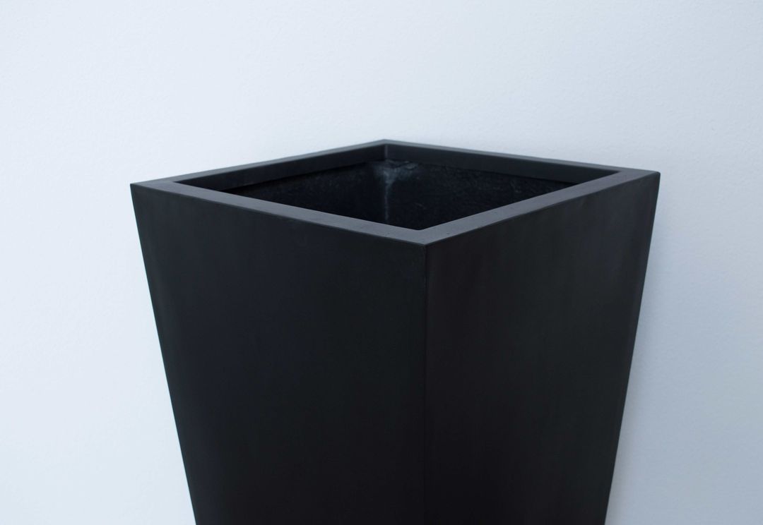 Oberer Teil eines konischen Pflanzkübels aus Fiberglas in Schwarz-matt mit quadratischer Öffnung vor heller Wand