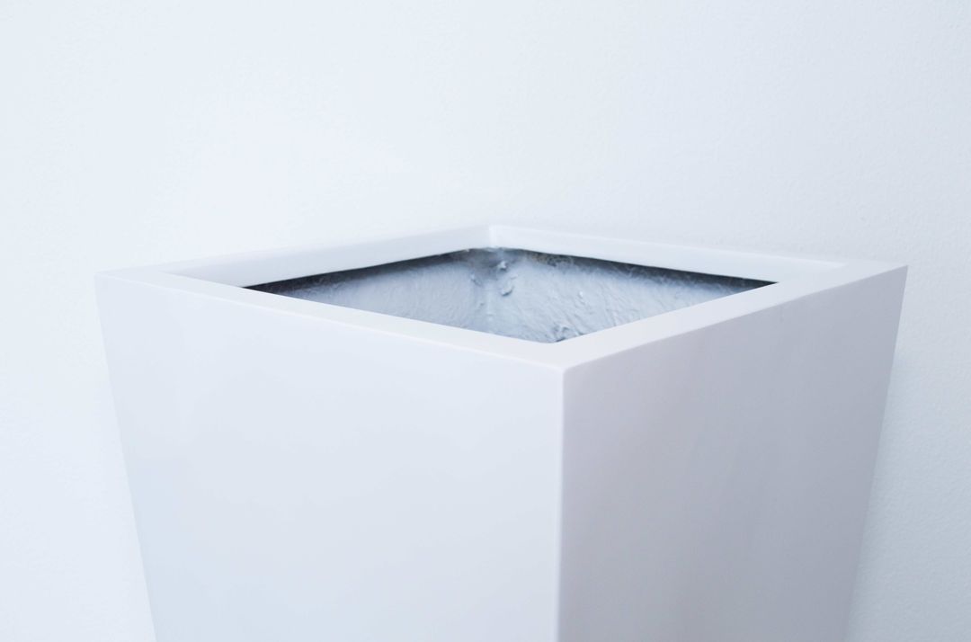 Weißer Pflanzkübel aus Fiberglas als hohe konische Säule vor weißer Wand auf braunem Fliesenboden
