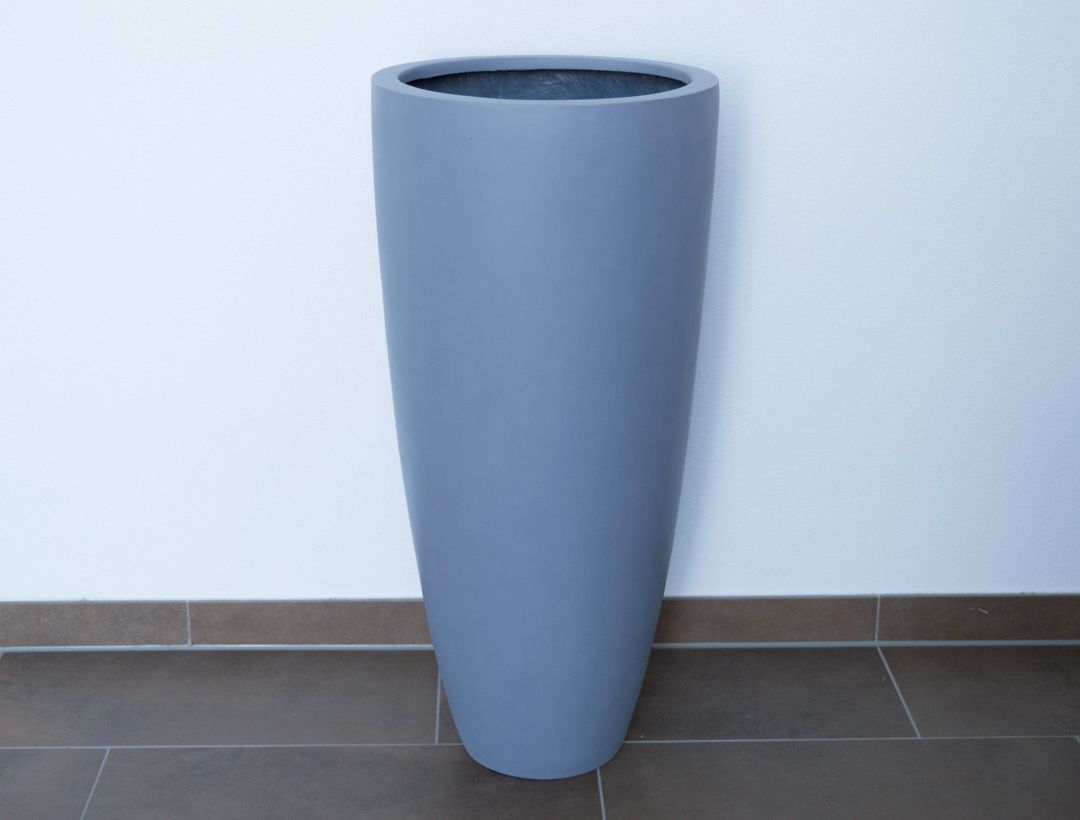 Hoher grauer Pflanzkübel aus Fiberglas in rund konischer Form ohne Bepflanzung auf braunem Fliesenboden vor weißer Wand