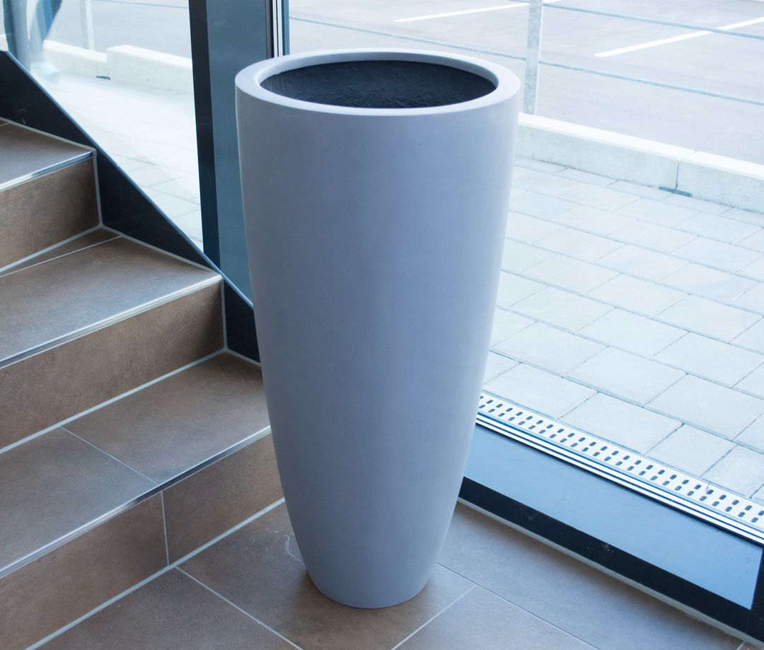 Grauer runder konischer Pflanzkübel aus Fiberglas ohne Pflanze neben einer Treppe vor einer Glasfront