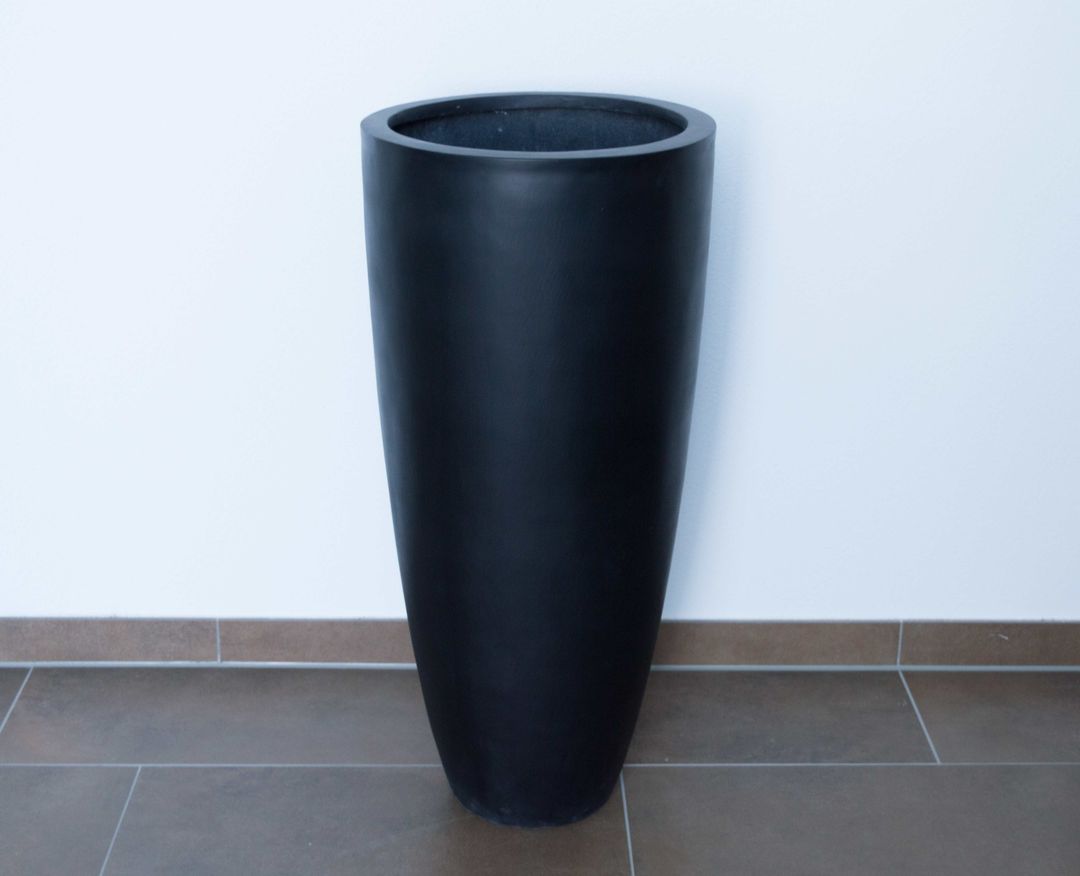 Hoher, runder konischer Pflanzkübel aus Fiberglas in Schwarz-matt, ohne Pflanze vor weißer Wand auf braunem Fliesenboden
