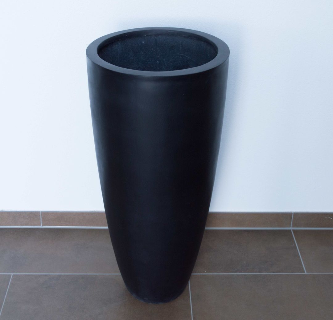 Schwarz-matter, hoher runder konischer Pflanzkübel aus Fiberglas vor weißer Wand auf braunem Fliesenboden