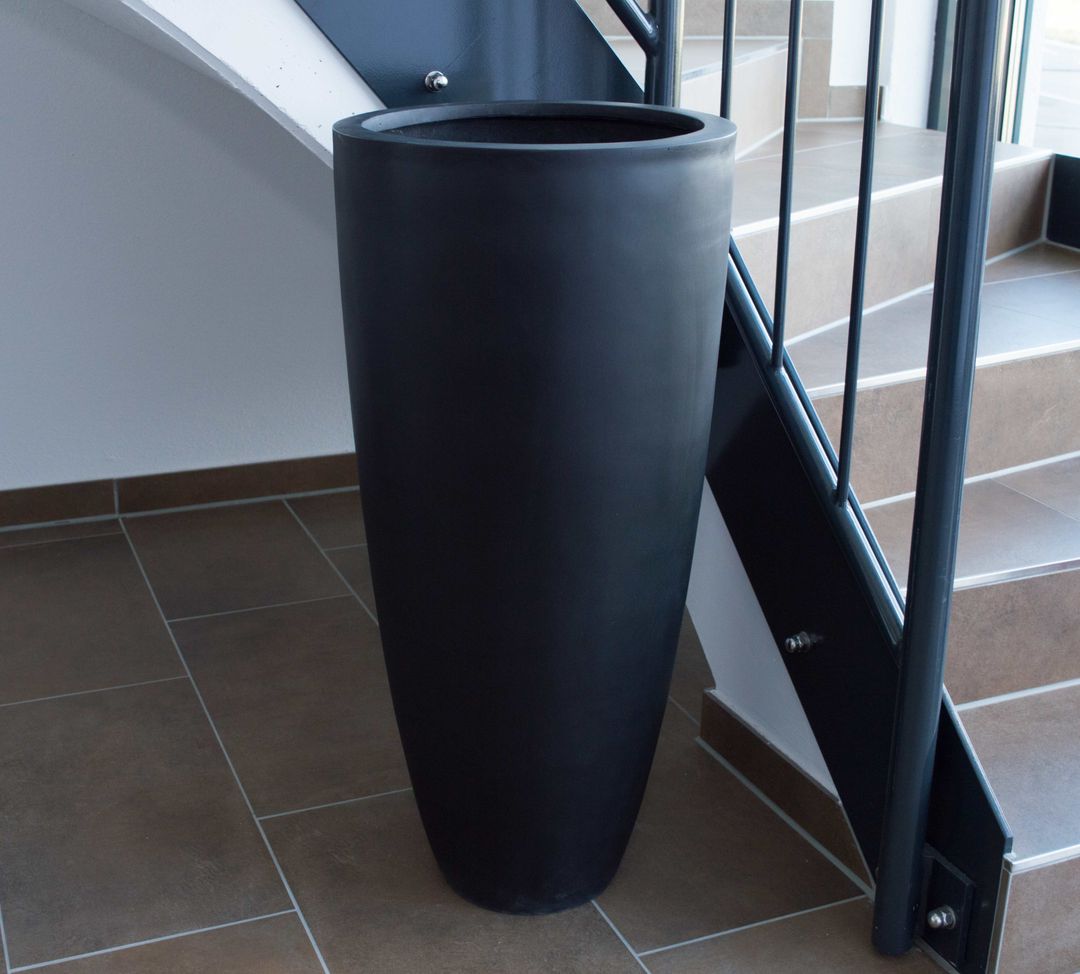 Schwarz-matter, rund konischer Pflanzkübel aus Fiberglas ohne Bepflanzung neben einer Treppe auf braunen Fliesen