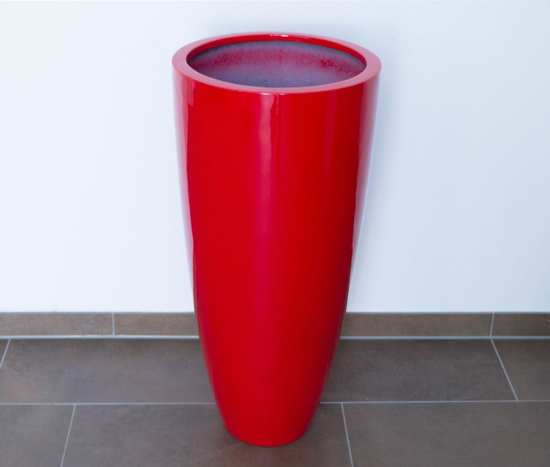 Hochglanz roter, rund konischer Pflanzkübel aus Fiberglas ohne Bepflanzung vor weißer Wand auf braunem Fliesenboden