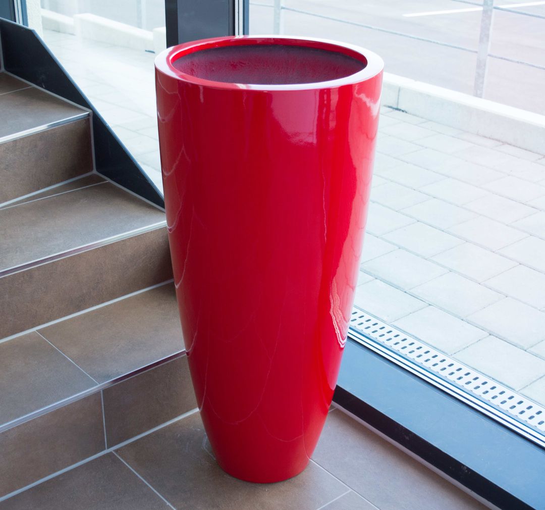 Hochglanz roter, rund konischer Pflanzkübel aus Fiberglas ohne Bepflanzung neben einer Treppe vor einer Glasfront