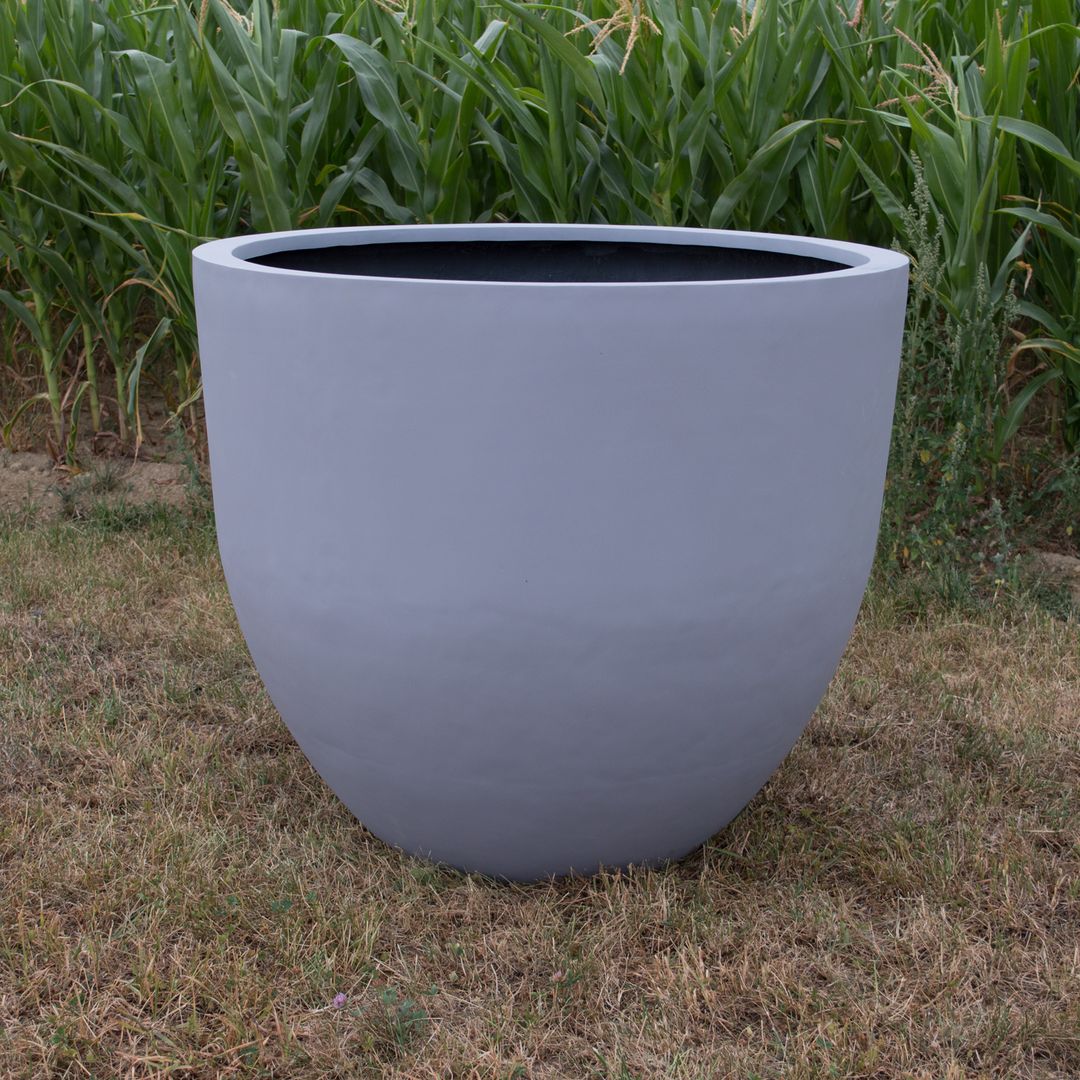 Großer grauer runder Pflanzkübel aus Fiberglas, unbepflanzt auf trockenem Rasen vor einem grünen Maisfeld