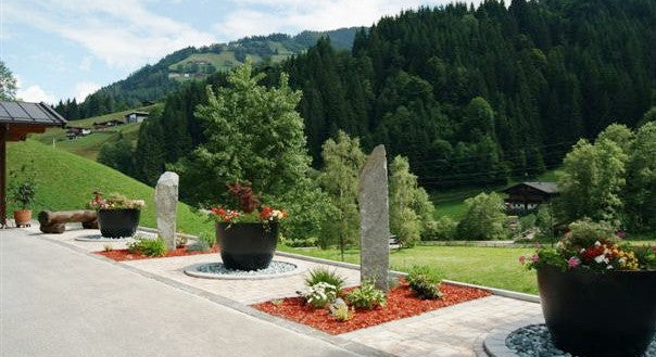 Schwarz-matte runde konische Pflanzkübel mit Sommerblumen an einer gepflasterten Einfahrt vor bewaldeten Bergen