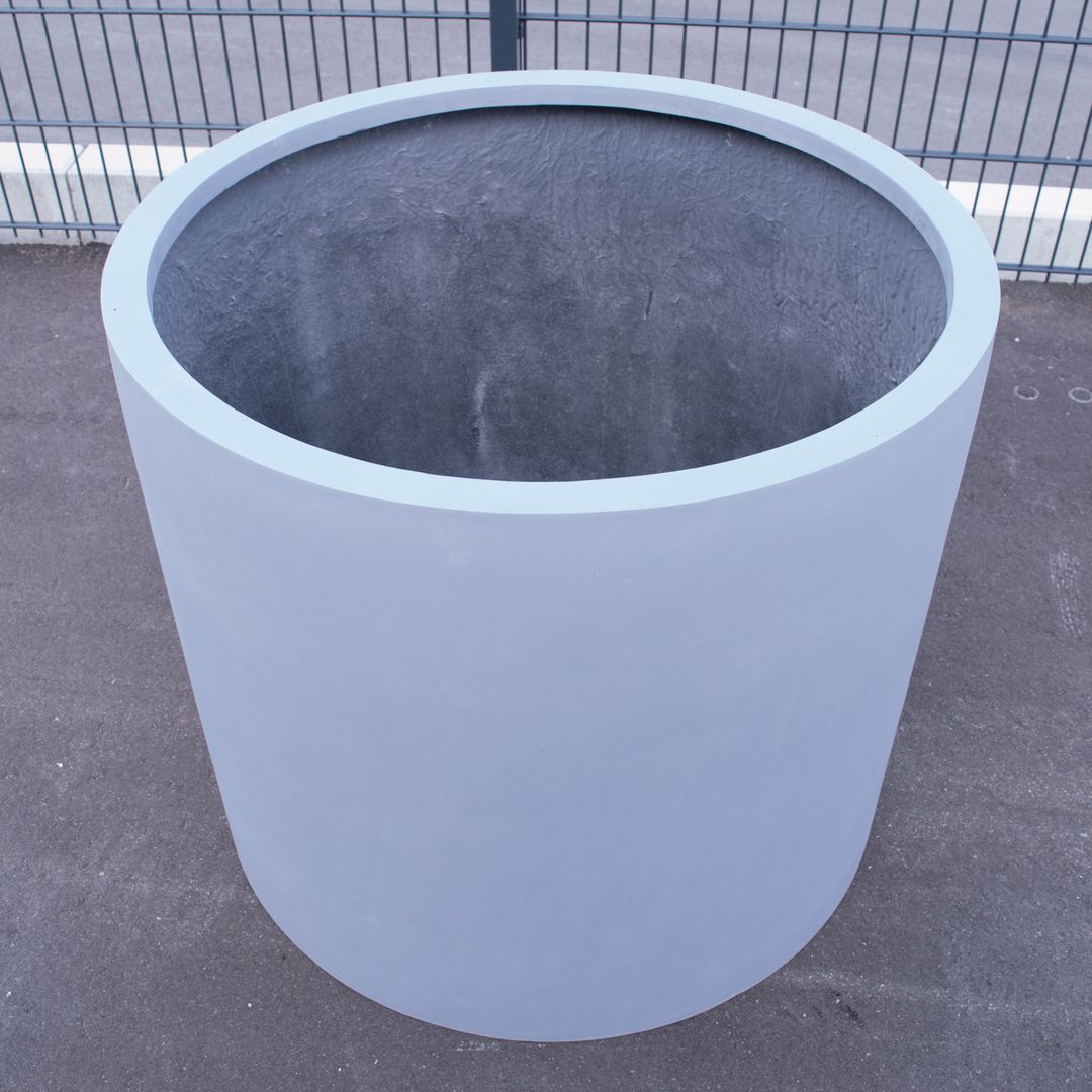 Leerer zylindrischer Pflanzkübel aus Fiberglas in Grau, schräg von oben fotografiert, auf Asphalt vor einem Metallzaun