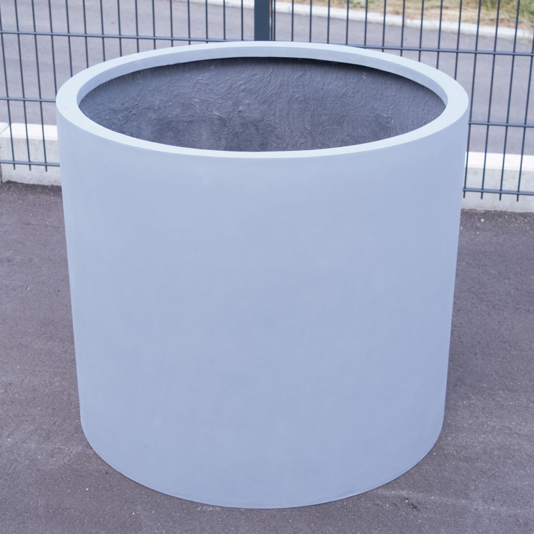 Grauer zylindrischer Pflanzkübel aus Fiberglas ohne Bepflanzung auf Asphalt vor einem Metallgitterzaun