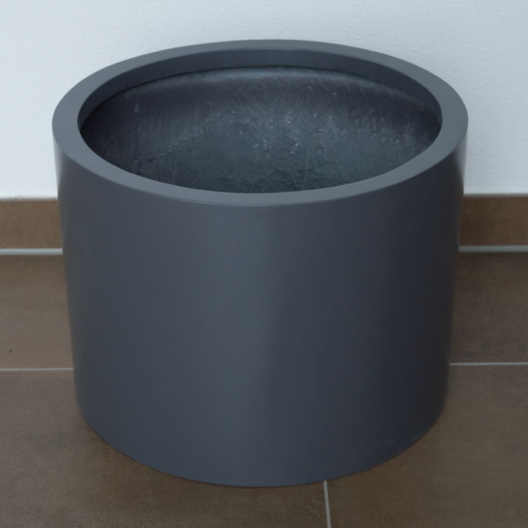 Zylindrischer Pflanzkübel aus Fiberglas in Anthrazit-Metallic, leer auf braunem Fliesenboden vor weißer Wand