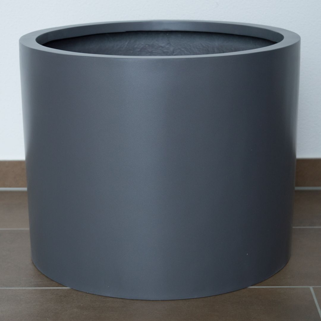 Zylindrischer Pflanzkübel aus Fiberglas in Anthrazit-Metallic, leer auf braunem Fliesenboden vor weißer Wand