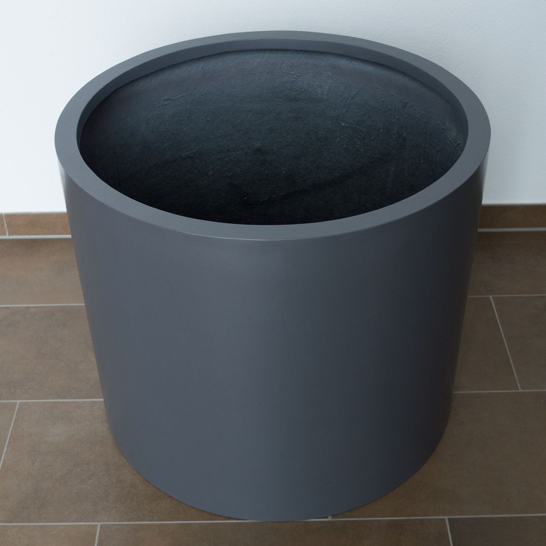 Leerer zylindrischer Pflanzkübel aus Fiberglas in Anthrazit-Metallic von schräg oben auf braunem Fliesenboden vor weißer Wand