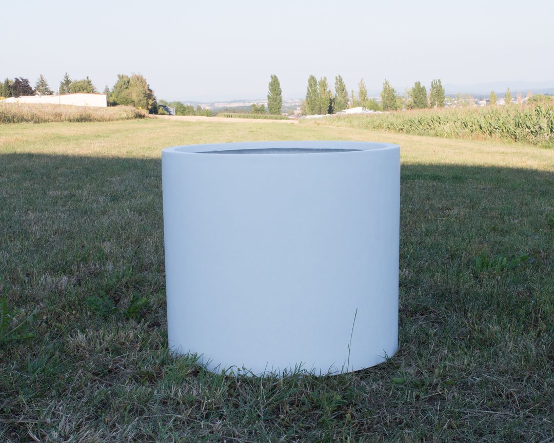 Leerer zylindrischer Pflanzkübel aus Fiberglas in Perlmuttweiß auf einer Wiese mit Bäumen und Feldern im Hintergrund