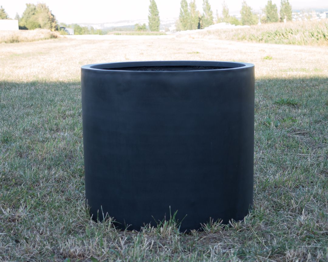 Schwarz-matter zylindrischer Pflanzkübel aus Fiberglas ohne Bepflanzung auf einer Wiese im Freien
