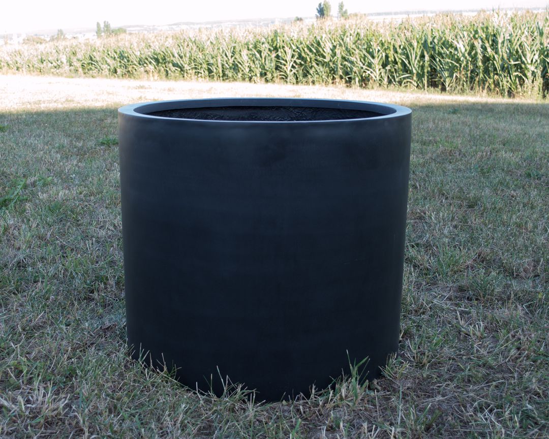 Zylindrischer Pflanzkübel aus Fiberglas in Schwarz-Matt, leer auf einer Wiese vor einem Maisfeld