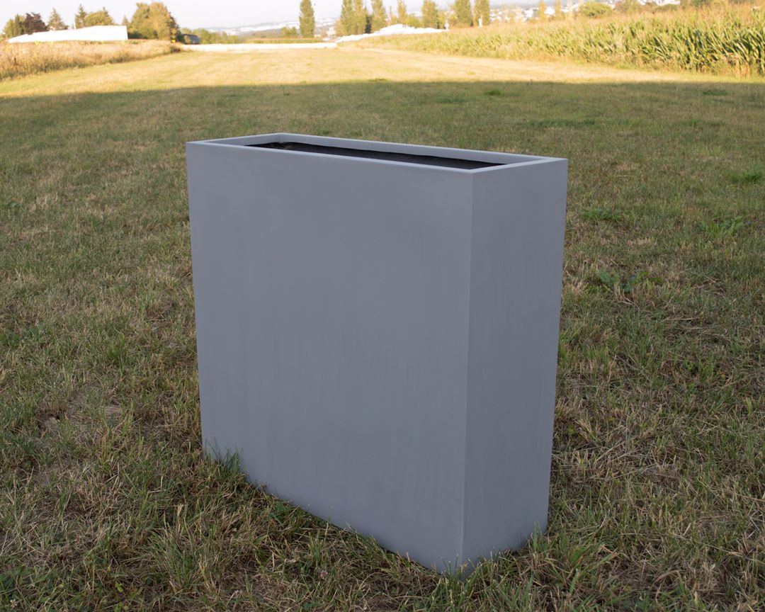 Grauer rechteckiger Pflanzkübel aus Fiberglas als Raumteiler, unbepflanzt auf einer Wiese im Freien