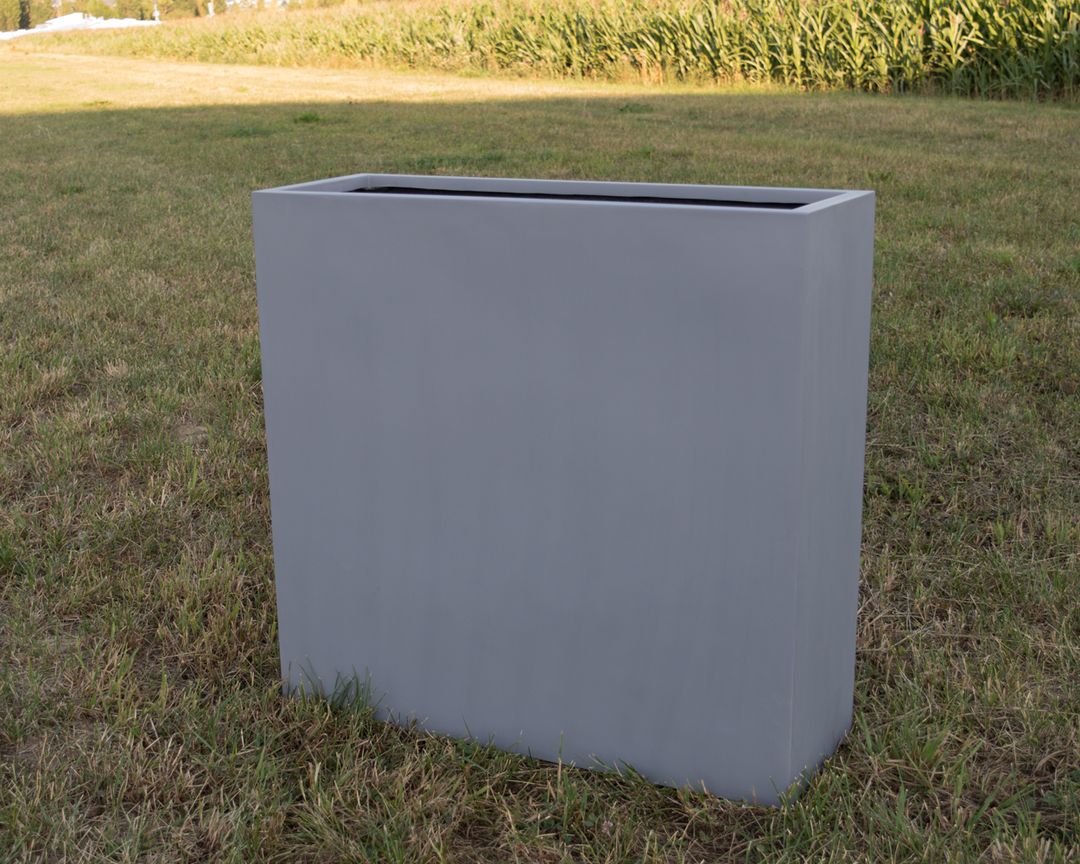 Schmaler, hoher Pflanzkübel aus Fiberglas in Grau, rechteckig und leer auf einer Wiese vor einem Maisfeld
