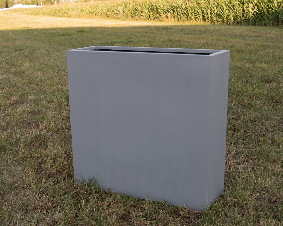 Schmaler, hoher Pflanzkübel aus Fiberglas in Grau, rechteckig und leer auf einer Wiese vor einem Maisfeld