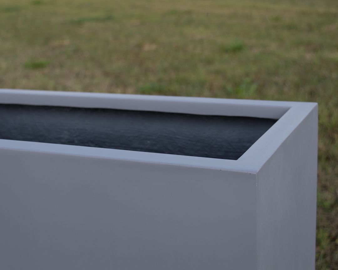 Nahaufnahme der oberen Ecke eines grauen rechteckigen Pflanzkübels aus Fiberglas mit dunklem Innenraum vor einer Rasenfläche