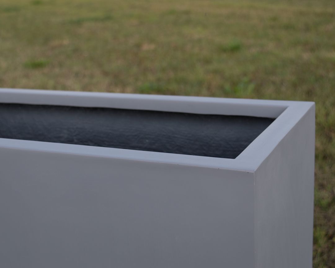 Nahaufnahme der oberen Ecke eines grauen rechteckigen Pflanzkübels aus Fiberglas mit dunklem Innenraum vor einer Rasenfläche