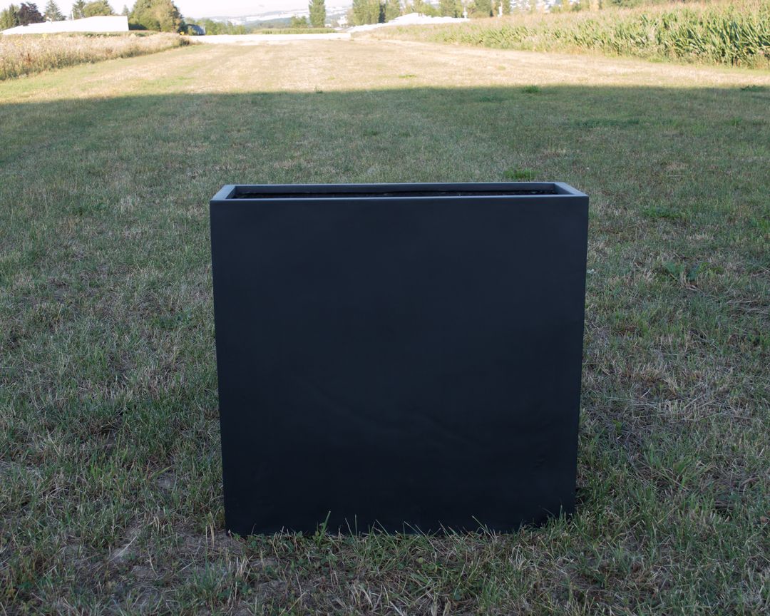 Rechteckiger Pflanzkübel aus Fiberglas in Schwarz-matt ohne Bepflanzung auf einer Wiese vor Feldern