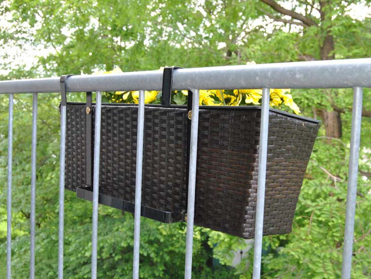 Rechteckiger Pflanzkübel aus Polyrattan in Coffee Braun mit gelben Blumen, am Balkongeländer vor grünen Bäumen eingehängt