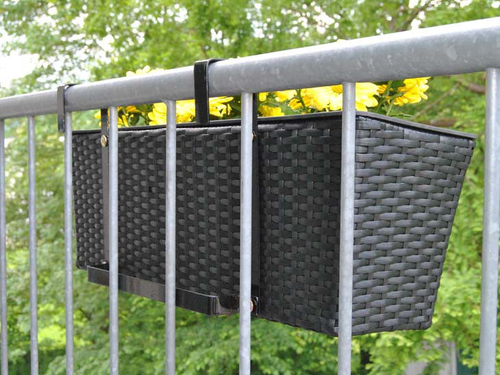 Schwarzer rechteckiger Pflanzkasten aus Polyrattan mit gelben Blumen, an einem Balkongeländer aus Metall eingehängt